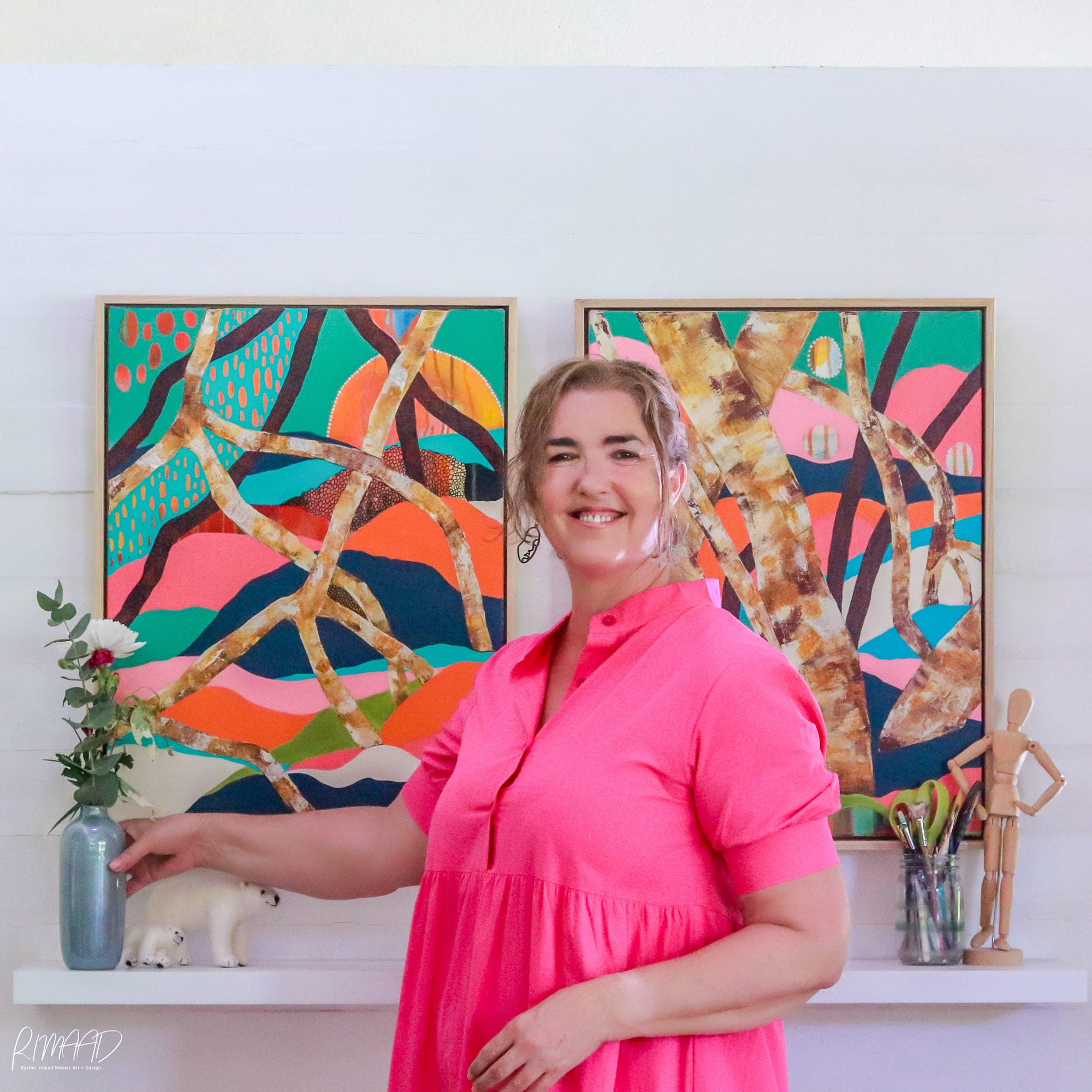 This is an image of an artist and woman standing in front of two original artworks. These artworks show coastal mangroves. These paintings are bright with pinks and greens, blues and yellow and orange. The are colourful and fun loving