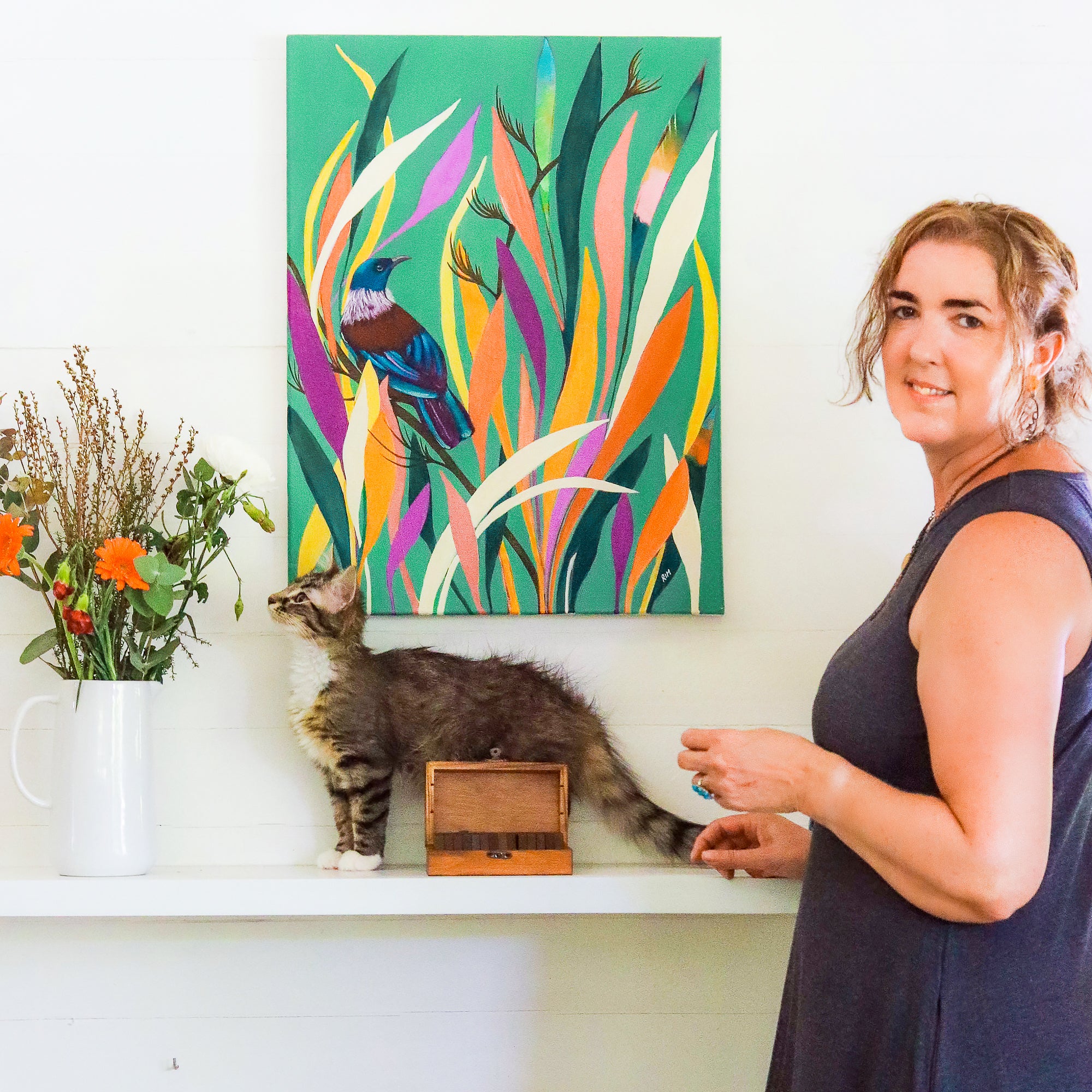 This is an image of a woman, standing in front of a painting with a Tui bird, and there is a small kitten and a vase of flowers.  